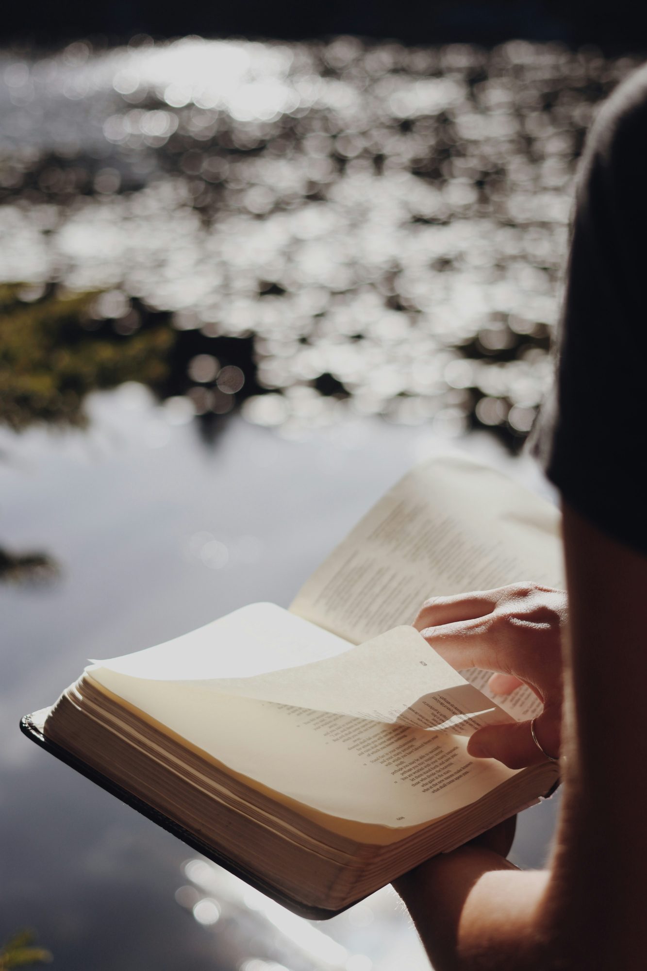 A person turning the pages of a Bible beside sunlit water